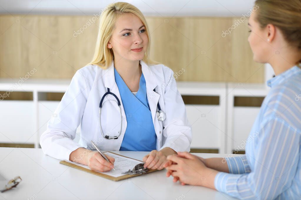 Woman doctor and patient sitting and talking at medical examination in hospital or clinic. Blonde therapist cheerfully smiling. Medicine and healthcare concept