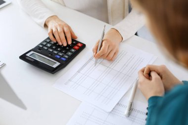 Muhasebeci mali durumu kontrol ediyor ya da vergi formları için hesap makinesi geliri ile sayıyor, eller yakın çekim. İş kadını ofisteki masada iş arkadaşıyla oturuyor ve çalışıyor. Denetim kavramı