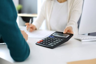 Muhasebeci mali durumu kontrol ediyor ya da vergi formları için hesap makinesi geliri ile sayıyor, eller yakın çekim. İş kadını ofisteki masada iş arkadaşıyla oturuyor ve çalışıyor. Denetim kavramı