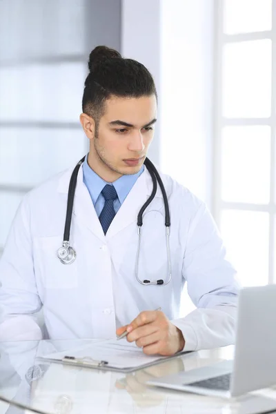 薬歴の記録を記入しながら、ラップトップコンピュータを使用してアラブの医師の男は、医療事務所や診療所のガラスデスクでフォーム。医学と医療の概念 — ストック写真