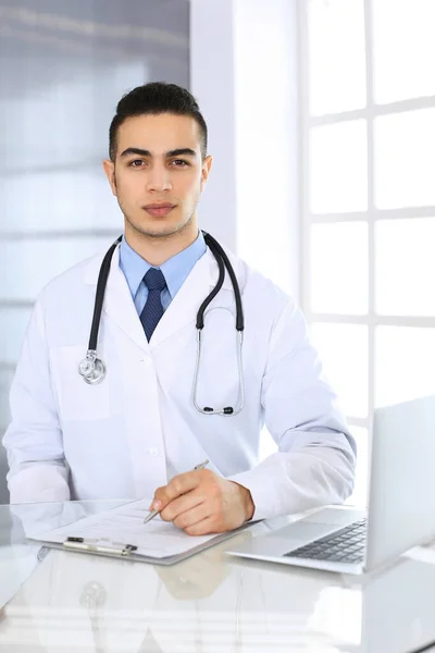Arabski lekarz korzystający z laptopa podczas wypełniania formularza historii leków w szklanym biurku w gabinecie medycznym lub klinice. Koncepcja medycyny i opieki zdrowotnej — Zdjęcie stockowe