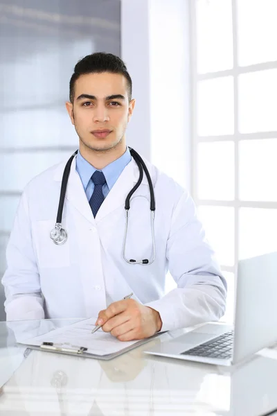 Medico arabo che utilizza il computer portatile durante la compilazione di record di farmaci modulo presso la scrivania di vetro in studio medico o in clinica. Medicina e concetto di assistenza sanitaria — Foto Stock