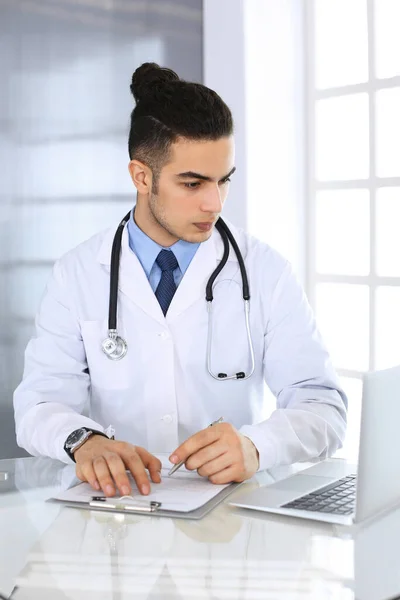 아랍 의사가 의료 사무실이나 진료소의 유리 책상에서 약물 기록을 작성하면서 노트북 컴퓨터를 사용하고 있다. 의학 과 건강 관리 개념 — 스톡 사진