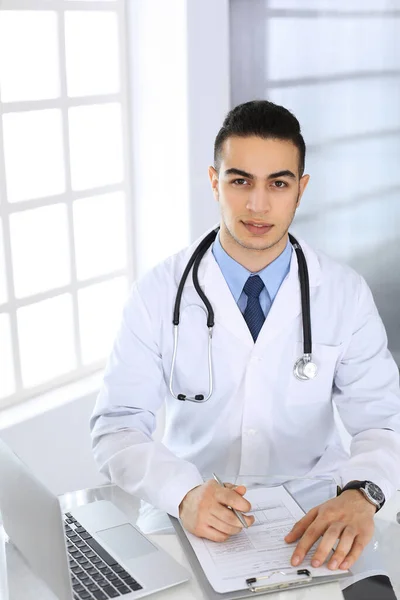 Arab lékař muž pomocí notebooku při vyplňování záznamů z historie léků formulář na skleněném stole v lékařské ordinaci nebo na klinice. Medicína a koncept zdravotní péče — Stock fotografie