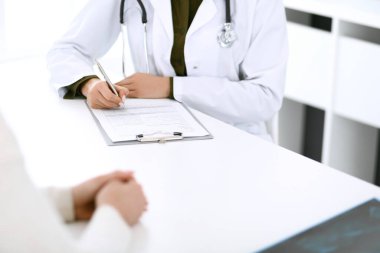 Kadın doktor ve hasta hastane muayenehanesinde oturuyor ve konuşuyor, yakın plan. İlaç kayıtlarını dolduran bir terapist. Tıp ve sağlık hizmetleri kavramı