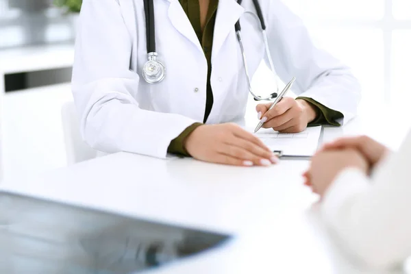 Kadın doktor ve hasta hastane muayenehanesinde oturuyor ve konuşuyor, yakın plan. İlaç kayıtlarını dolduran bir terapist. Tıp ve sağlık hizmetleri kavramı