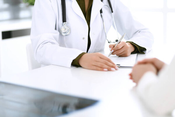 Woman doctor and patient sitting and talking at medical examination at hospital office, close-up. Therapist filling up medication history records. Medicine and healthcare concept