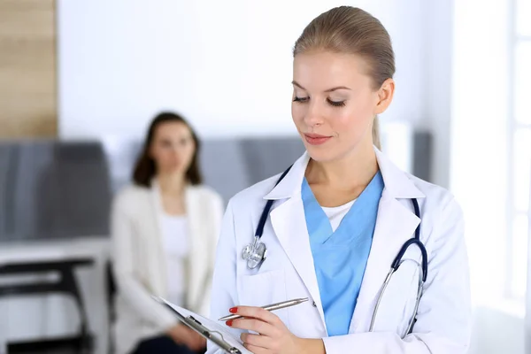 Läkare kvinna fylla upp medicinhistoria rekord när du står i akut sjukhus kontor med patienten i kö i bakgrunden. Läkare på jobbet, porträttfotografering. Medicin och hälsovård — Stockfoto