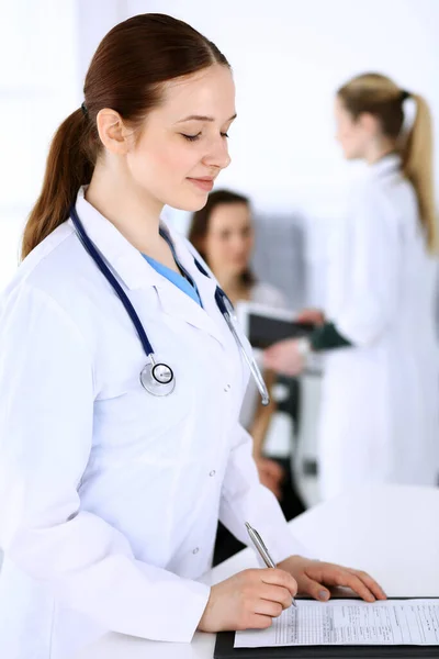 Läkare kvinna på jobbet med patient och kollega på bakgrunden. Läkare fylla upp medicinska dokument eller recept när du står på sjukhus receptionen. Uppgifter inom medicin och hälsovård — Stockfoto