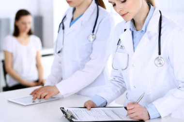 Hastanede çalışan bir grup doktor ve arka planda sırada bekleyen bir hasta. Doktor resepsiyonda beklerken tıbbi belge ya da reçete dolduruyor. Tıp ve sağlık verileri