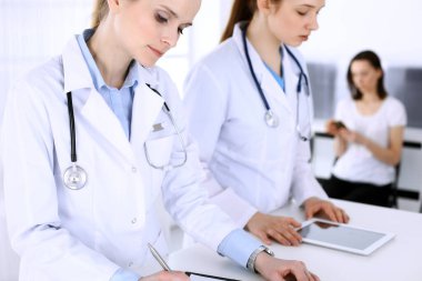 Hastanede çalışan bir grup doktor ve arka planda sırada bekleyen bir hasta. Doktor resepsiyonda beklerken tıbbi belge ya da reçete dolduruyor. Tıp ve sağlık verileri