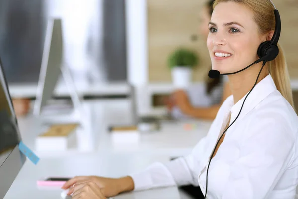 Donna d'affari bionda che utilizza auricolari per la comunicazione e la consulenza di persone presso l'ufficio del servizio clienti. Call center. Gruppo di operatori al lavoro sullo sfondo — Foto Stock