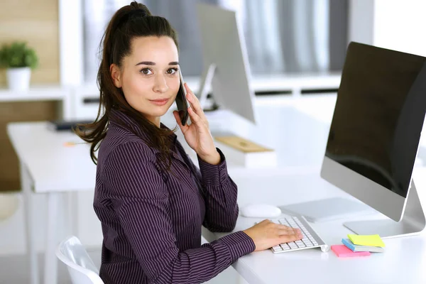 Business woman using phone and computer at workplace in modern office. Brunette secretary or female lawyer smiling and looks happy. Working for pleasure and success