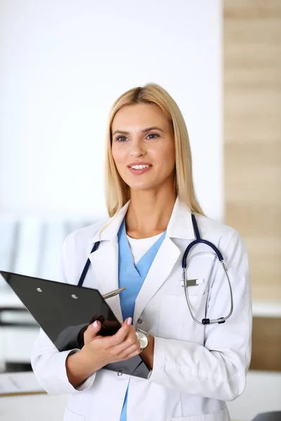 Doctora mujer en el trabajo en el hospital emocionada y feliz de su profesión. Médico rubio controla los registros de la historia de la medicación y los resultados del examen mientras usa portapapeles. Concepto de medicina y salud — Foto de Stock