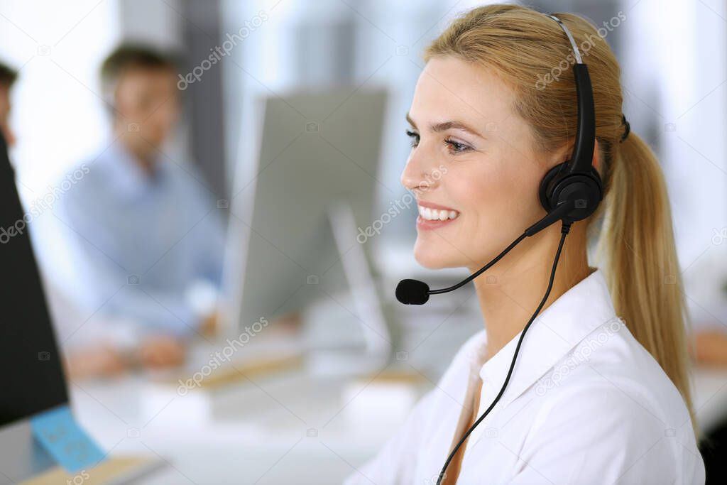 Blonde business woman using headset for communication and consulting people at customer service office. Call center. Group of operators at work at the background