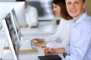 Modern ofiste neşeli gülümseyen iş adamı vesikalığı. Arka planda çeşitli iş arkadaşlarıyla otururken bilgisayar kullanan sıradan giyinmiş bir girişimci. Çoklu etnik çalışma