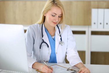 Hastanedeki ya da klinikteki masada oturan doktor kadın. Sarışın, neşeli doktor tıbbi kayıtları dolduruyor. Veriler ve tıp ve sağlık hizmetlerindeki en iyi hizmet