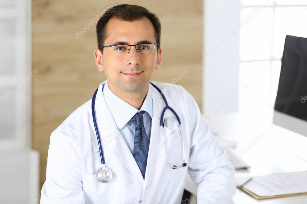 Doctor man sitting at the desk at his working place and smiling at camera. Perfect medical service in clinic. Happy future of medicine and healthcare