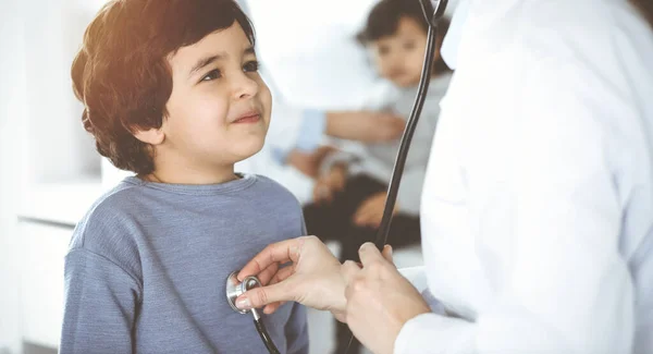 Médico-mujer examinando a un niño paciente por estetoscopio en Sunny Clinik. Lindo chico árabe y su hermano en la cita médica —  Fotos de Stock