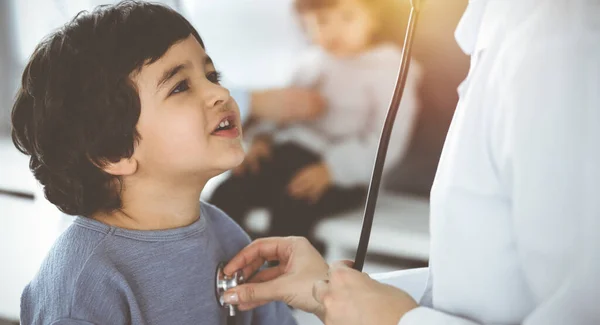 Médico-mujer examinando a un niño paciente por estetoscopio en Sunny Clinik. Lindo chico árabe y su hermano en la cita médica — Foto de Stock