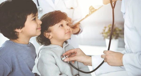 Médico-mujer examinando a un niño paciente por estetoscopio en Sunny Clinik. Lindo chico árabe y su hermano en la cita médica —  Fotos de Stock