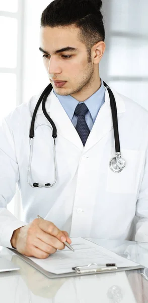 Arab doctor man is using laptop computer while filling up medication history records form at the glass desk in r clinic. Medicine concept — Stock Photo, Image