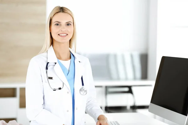 Médica loira está de pé em seu local de trabalho perto do computador desktop. Mulher-médico está animado e feliz de sua profissão. Conceito de medicina — Fotografia de Stock