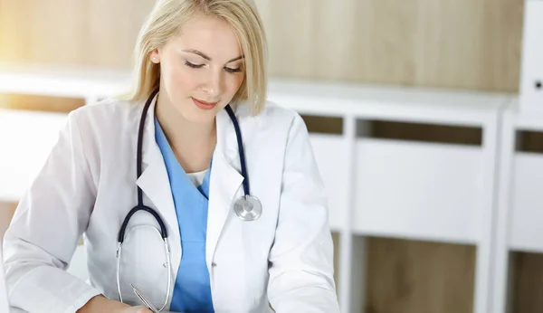 Mujer-médico en el trabajo mientras está sentado en el escritorio en la clínica soleada. Rubia alegre médico llenar la historia de la medicación formulario de registro — Foto de Stock