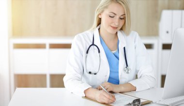 Güneşli klinikte masa başında oturan kadın doktor. Sarışın, neşeli doktor ilaç geçmişi formunu dolduruyor.