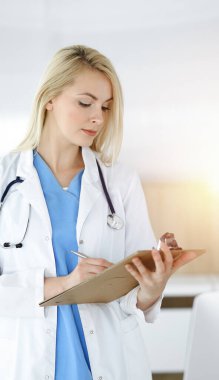 Kadın doktor güneşli klinikte duruyor ve ilaç geçmişi formunu dolduruyor. Tıptaki veriler