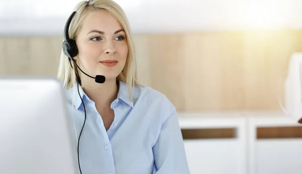 Blonde business woman sitting and communicated by headset in call center in sunny office. Concept of telesales business