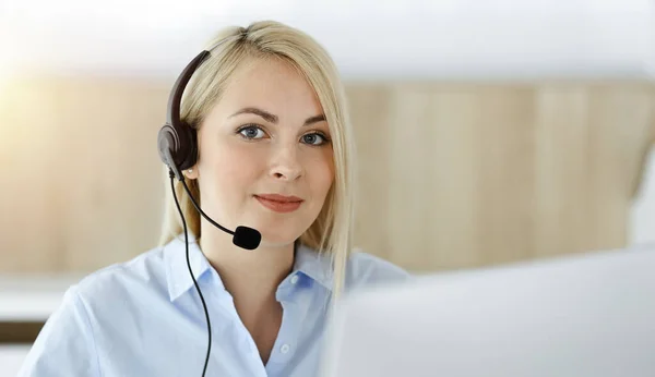 Mujer rubia de negocios sentada y comunicada por auriculares en call center en oficina soleada. Concepto de negocio de televentas — Foto de Stock