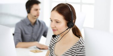 İnternette müşterilerle konuşurken kulaklık ve bilgisayar kullanan sıradan giyinmiş genç bir kadın. Bir grup operatör iş başında. Çağrı merkezi, iş konsepti