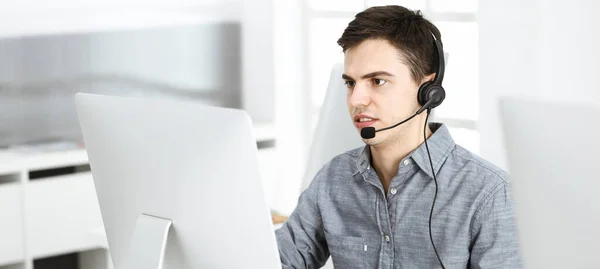 Hombre joven vestido casual usando auriculares y computadora mientras habla con los clientes en línea. Call center, concepto de negocio —  Fotos de Stock