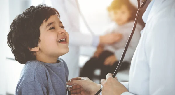 Médico-mujer examinando a un niño paciente por estetoscopio en Sunny Clinik. Lindo chico árabe y su hermano en la cita médica — Foto de Stock