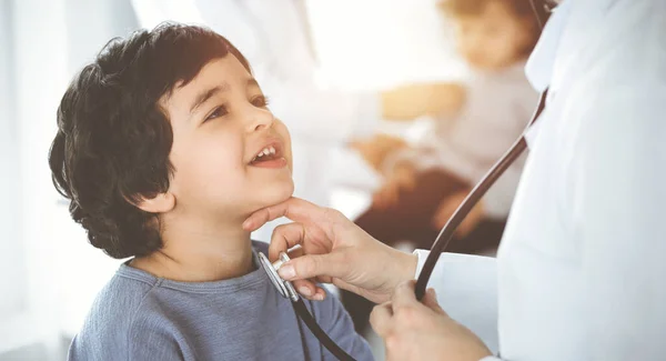 Médico-mujer examinando a un niño paciente por estetoscopio en Sunny Clinik. Lindo chico árabe y su hermano en la cita médica —  Fotos de Stock