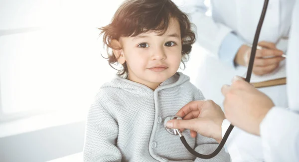 Ärztin untersucht eine Kinderpatientin per Stethoskop in einer sonnigen Klinik. Niedliches arabisches Kleinkind beim Arzttermin — Stockfoto