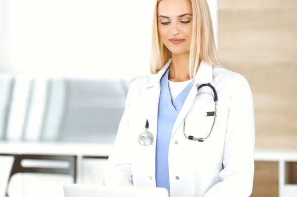 La médica rubia está sonriendo mientras usa la computadora portátil. Mujer-médico en el trabajo en la clínica emocionada y feliz de su profesión. Concepto de medicina — Foto de Stock
