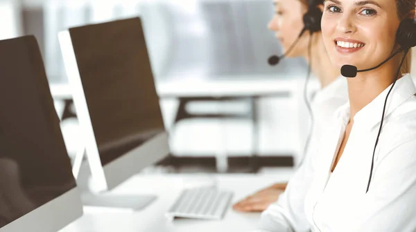 Mujer rubia representante de servicio al cliente y su colega están consultando a los clientes en línea utilizando auriculares. Centro de llamadas y concepto de gente de negocios — Foto de Stock