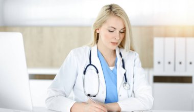 Güneşli klinikte masa başında oturan kadın doktor. Sarışın, neşeli doktor ilaç geçmişi formunu dolduruyor.