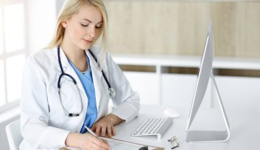 Güneşli klinikte masa başında oturan kadın doktor. Sarışın, neşeli doktor ilaç geçmişi formunu dolduruyor.