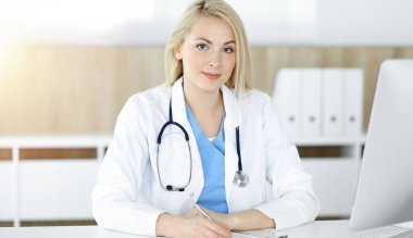 Güneşli klinikte masa başında oturan kadın doktor. Sarışın, neşeli doktor ilaç geçmişi formunu dolduruyor.