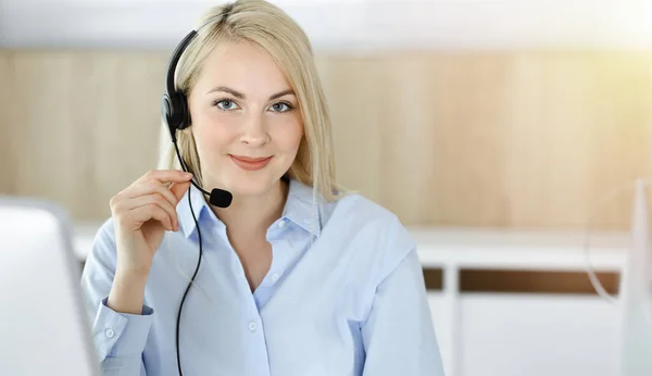 Mujer rubia de negocios sentada y comunicada por auriculares en call center en oficina soleada. Concepto de negocio de televentas — Foto de Stock