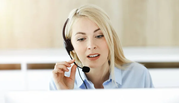 Donna d'affari bionda seduta e comunicata da auricolare in call center in ufficio soleggiato. Concetto di attività di televendita — Foto Stock