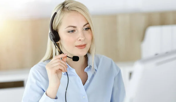 Blond affärskvinna sitter och kommuniceras med headset i call center i soligt kontor. Begreppet telehandel Stockbild