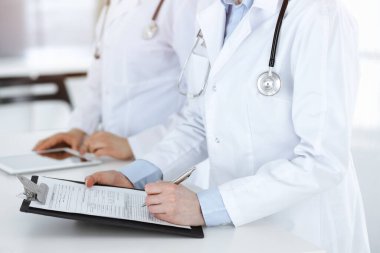 İş yerindeki bilinmeyen kadın doktorlar, arka planda hastaları var. Güneşli hastane resepsiyonunda beklerken tıbbi belgeleri dolduran kadın doktorlar, yakın plan. Sağlık hizmetleri