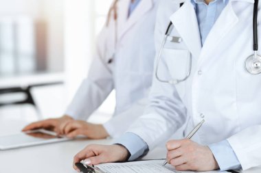 İş yerindeki bilinmeyen kadın doktorlar, arka planda hastaları var. Güneşli hastane resepsiyonunda beklerken tıbbi belgeleri dolduran kadın doktorlar, yakın plan. Sağlık hizmetleri