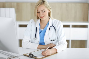 Hastanedeki ya da klinikteki masa başında oturan kadın doktor. Sarışın, neşeli doktor ilaç geçmişi formunu dolduruyor.