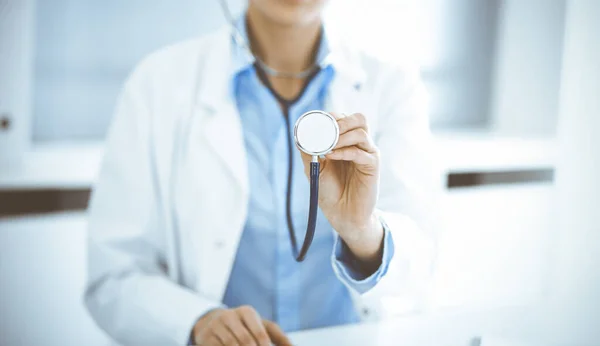 Okänd kvinna-läkare i blå blus håller stetoskop huvudet medan du sitter vid skrivbordet på kliniken, närbild. Läkare redo att undersöka och hjälpa patienten. Medicinskt koncept — Stockfoto