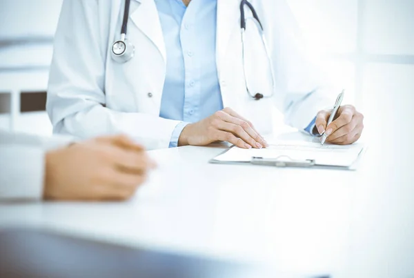 Mujer-médico desconocida y paciente sentada y hablando en el examen médico en la clínica, de cerca. El terapeuta que usa blusa azul está llenando el historial de medicamentos. Concepto de medicina — Foto de Stock